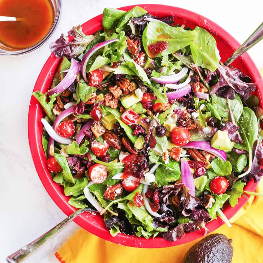 Salad tongs tucked into a big red bowl of chopped salad with a fresh avocado sitting next to the bowl alongside a cup of dressing. 