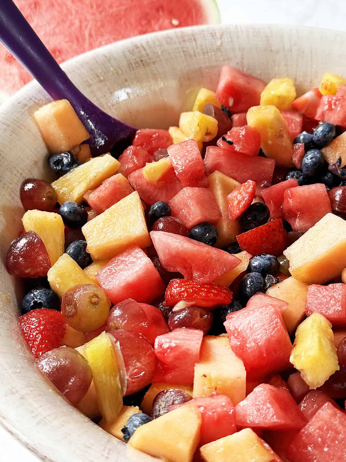 Bowl of fruit salad with a serving spoon stuck inside and a half of a watermelon in the background.