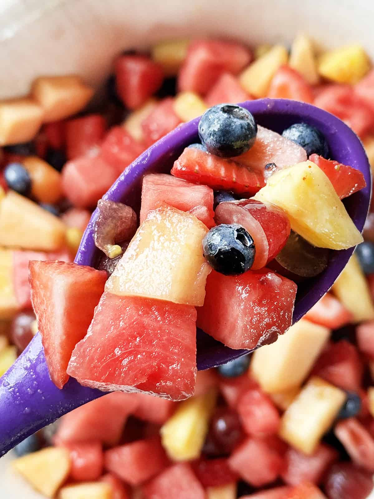 Serving spoon with fruit salad on it hovering over a serving bowl filled with the mixture.