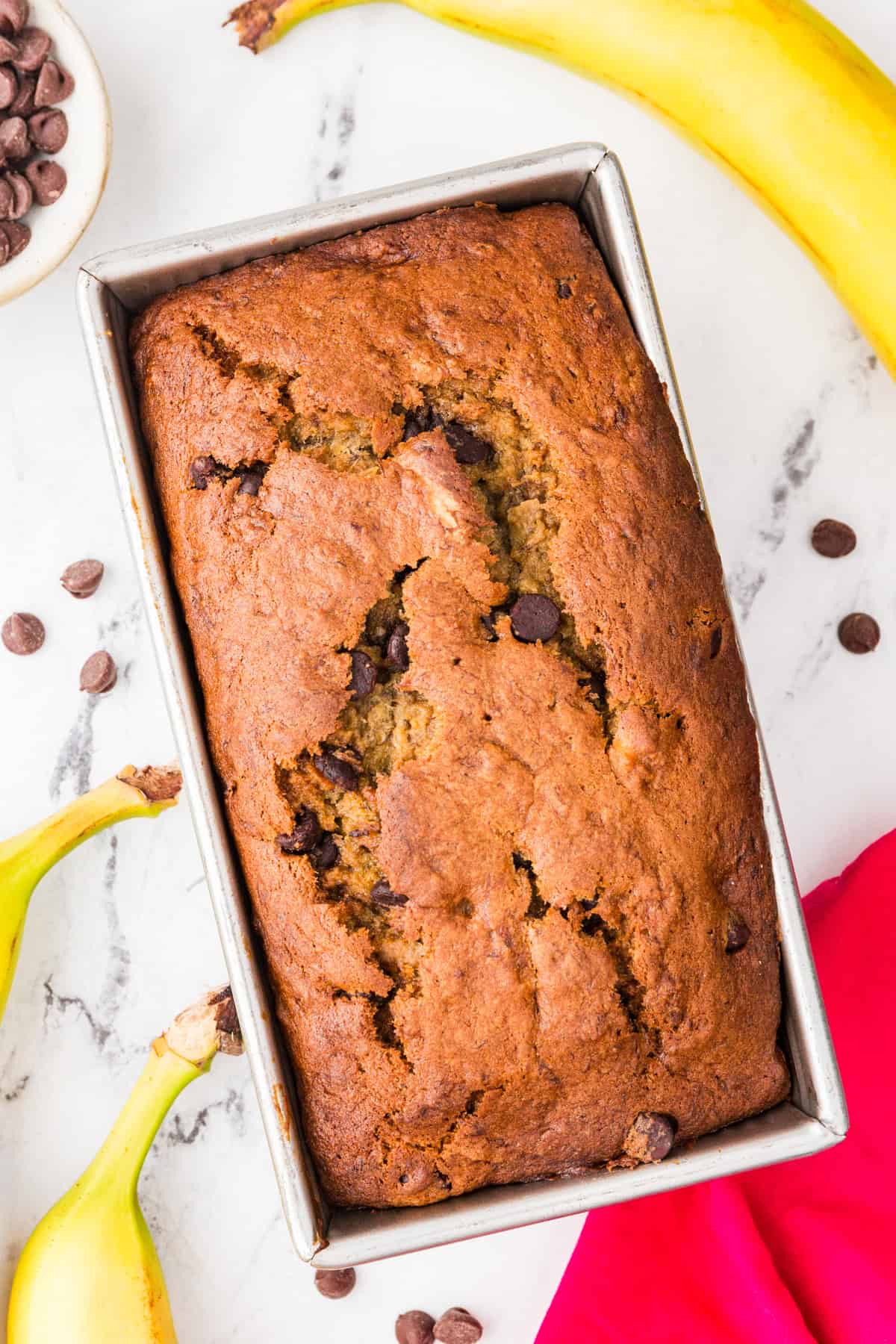 Top view of a baked loaf of banana bread with chocolate chips inside, and surrounded by bananas.