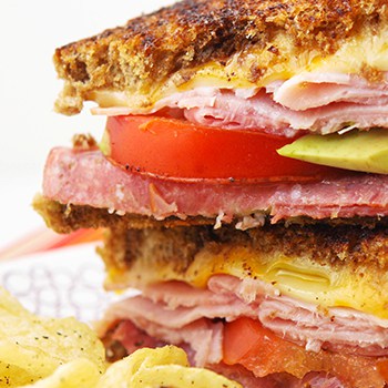 Close up shot of a leftover lunch meat grilled cheese sandwich cut in half and stacked on top of one another with chips on the side. 