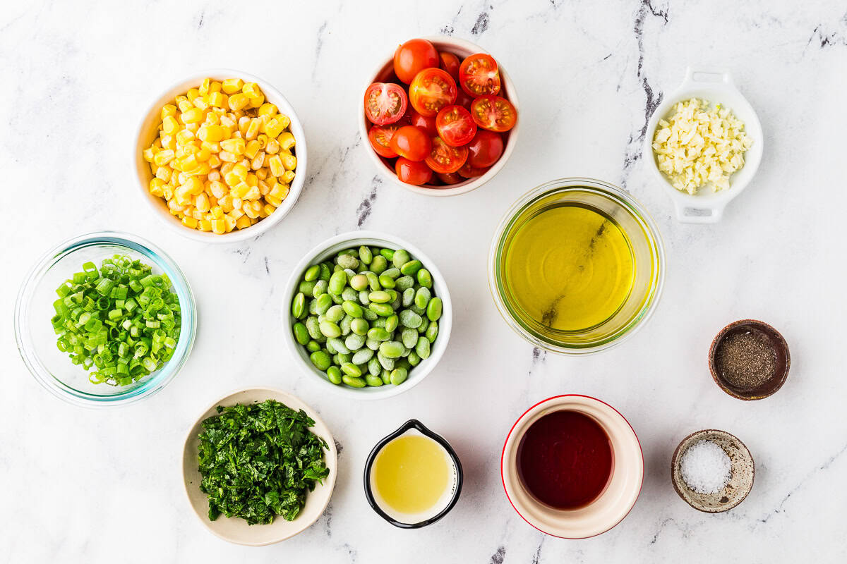 Ingredients for edamame tomato corn salad in individual bowls, lined up on a white counter.