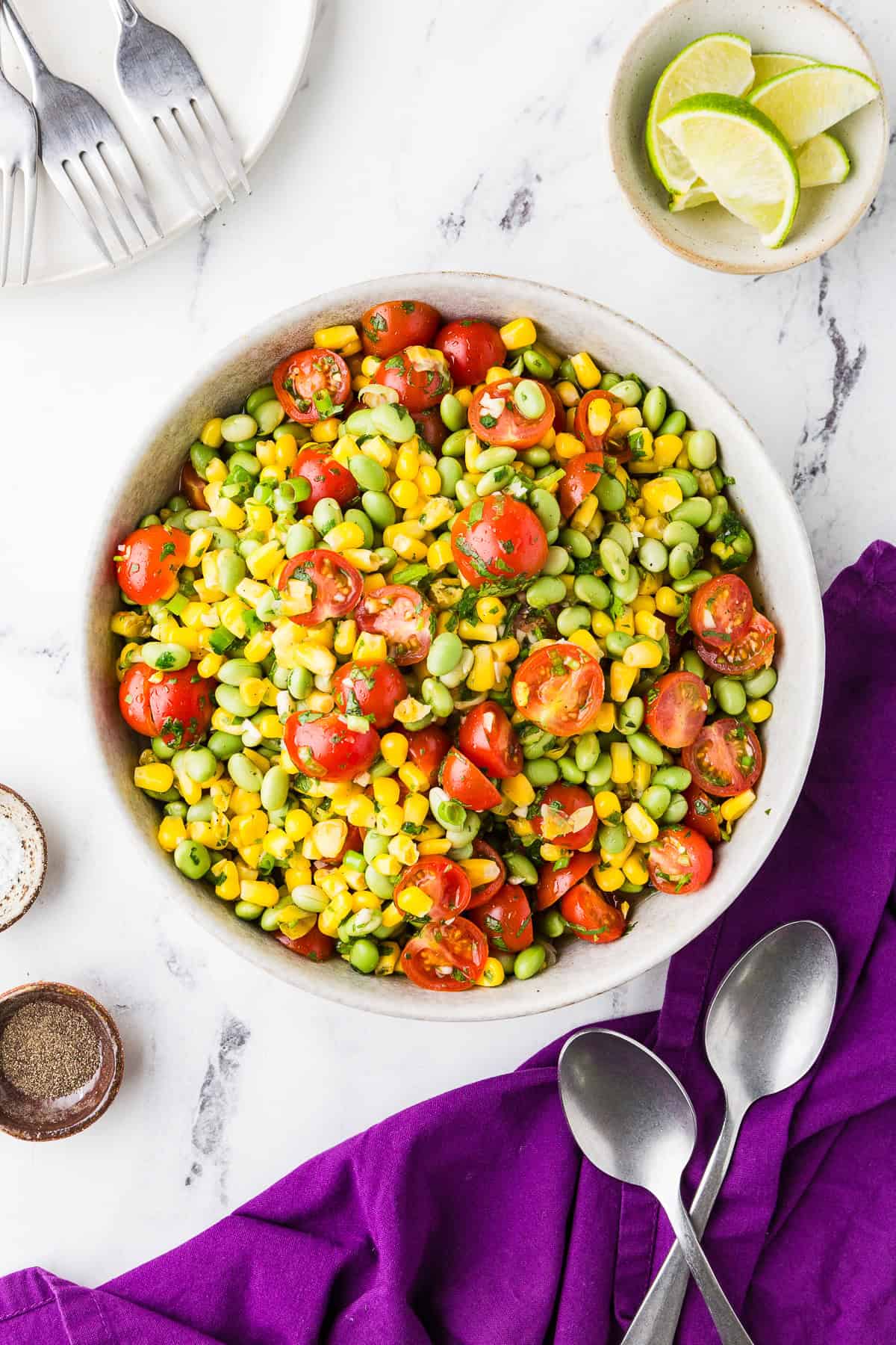 Top view of a serving bowl filled with edamame summer tomato salad next to spoons and lime wedges.