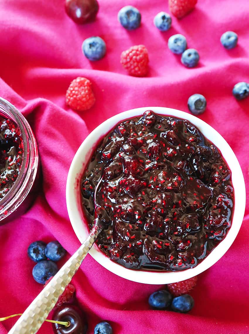 Delicious looking dark colored jam in a bowl, surrounded by berries and cherries on a placemat.