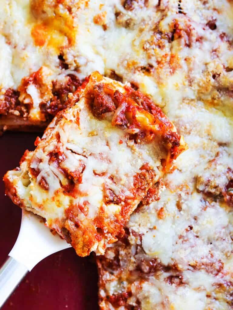 Spatula holding a piece of cheesy lasagna hovering over baking dish.