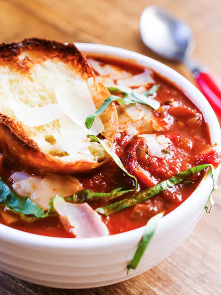 Roasted tomato soup served in a bowl with strips of basil garnished on top next to a piece of toast sitting on top of the bowl. 