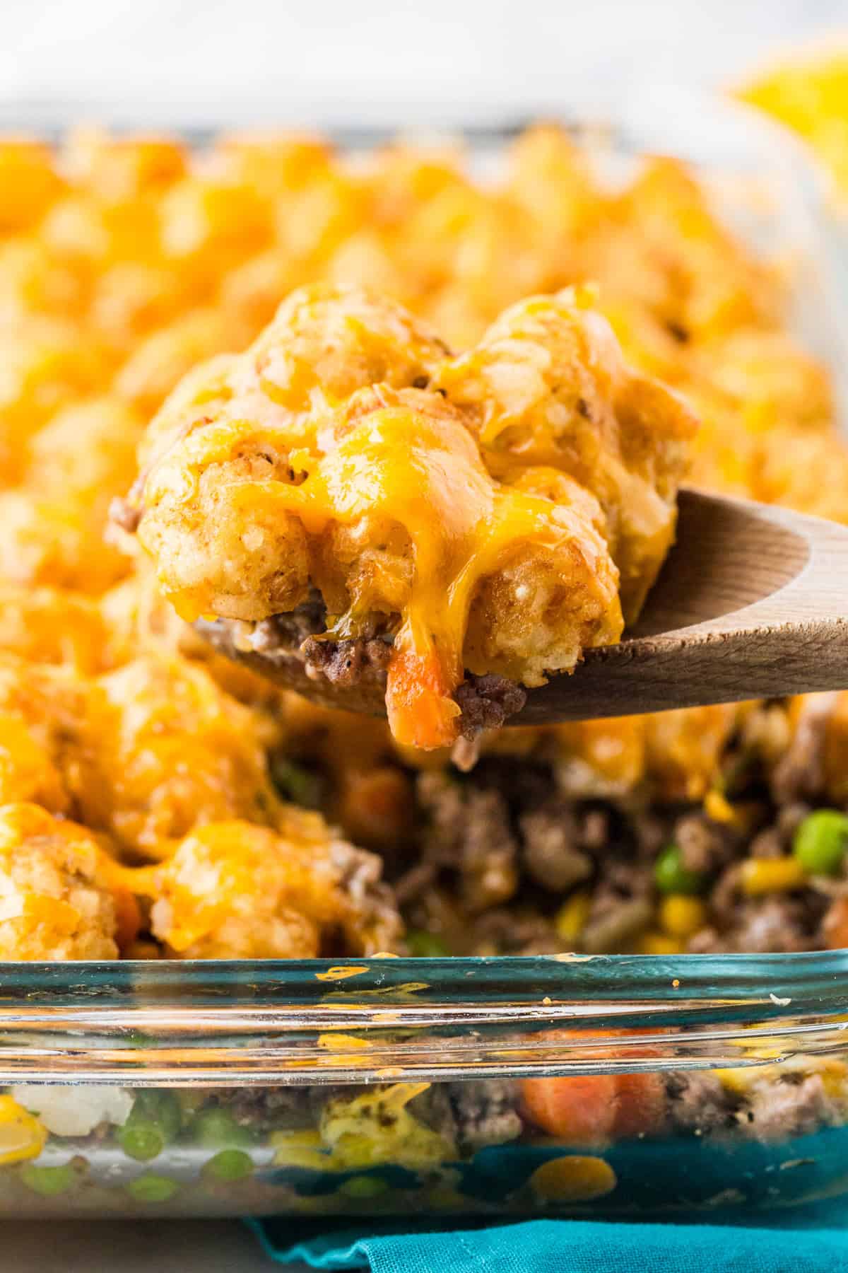 Wooden spoon lifting a spoonful of cheesy, meaty casserole topped with tater tots out of a baking dish.