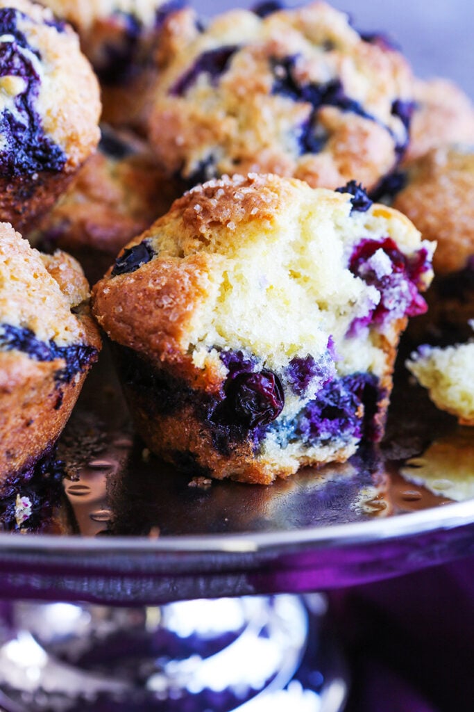 A pedestal full of homemade blueberry muffins with one split in half to see juicy blueberries inside. 