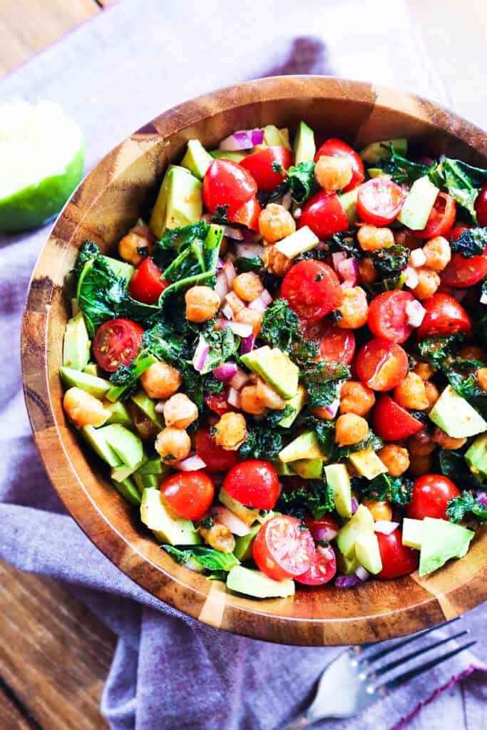 Bowl of kale salad with chickpeas and tomatoes, avocado and cucumbers on top. 