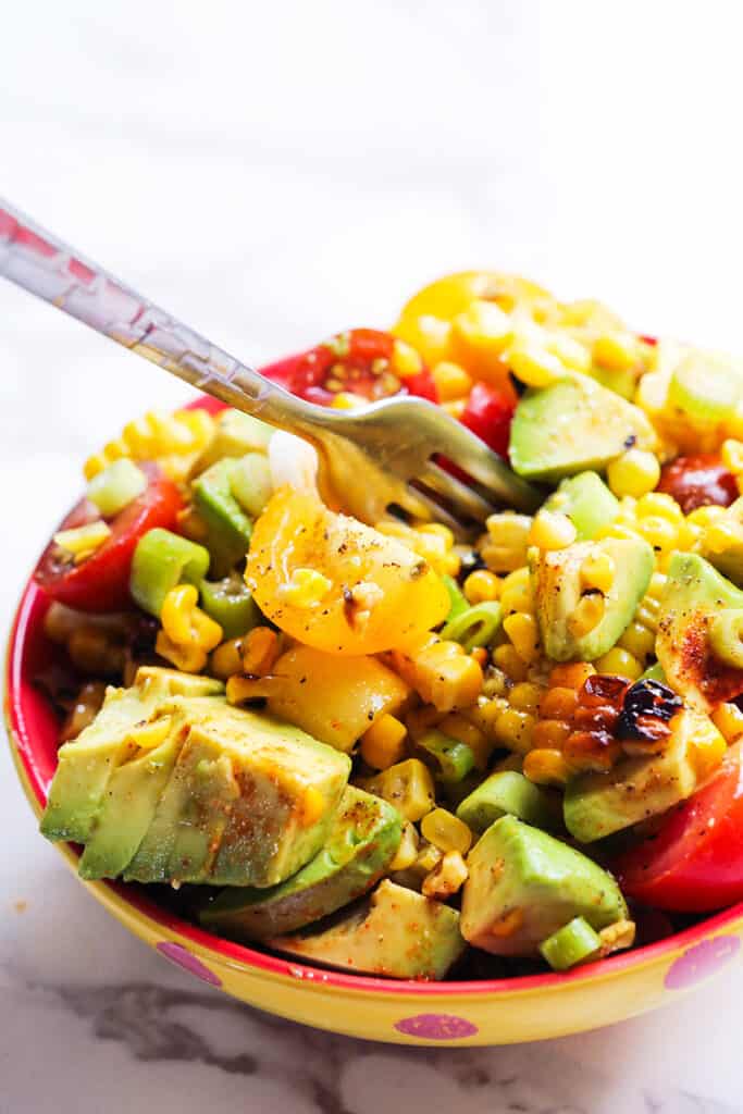 A fork stuck in a serving bowl of grilled corn and tomato salad with fresh avocado slices topping it. 