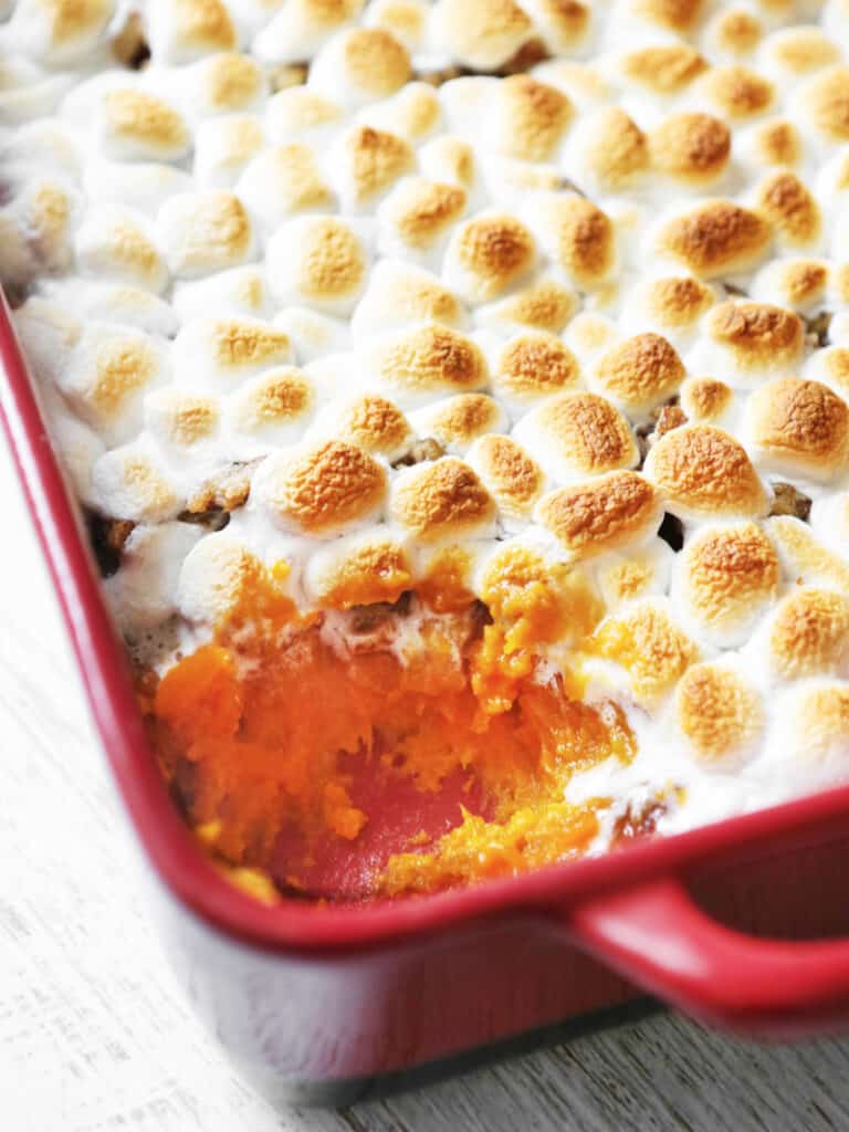 A scoop taken out of a casserole pan  filled with sweet potato casserole topped with melted marshmallows. 