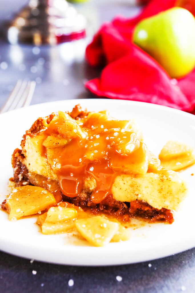 slice of salted caramel cheesecake with apples on top and caramel drizzled over the slice sitting on a plate. 