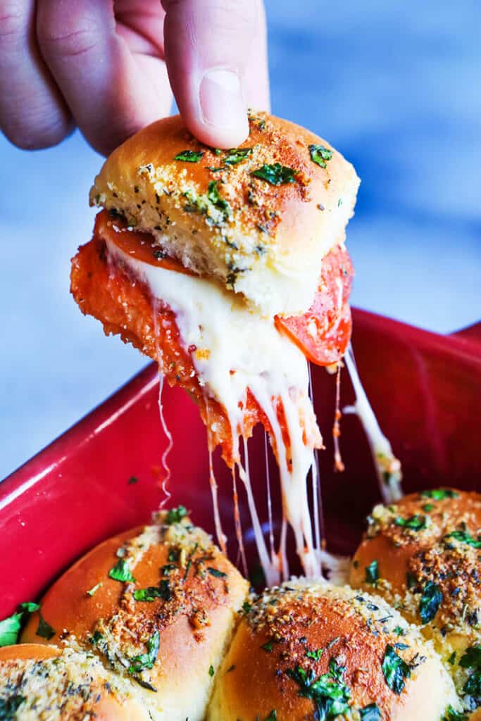 Fingers picking up a pizza slider out of a casserole dish with melted cheese hanging down. 