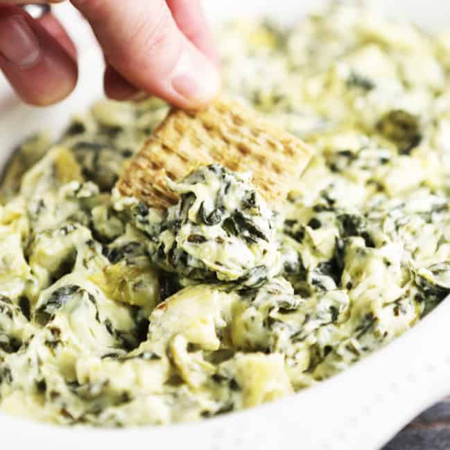 Hand using a cracker to scoop up hot spinach artichoke dip. 