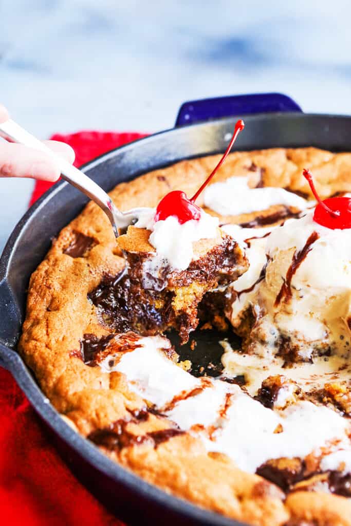 spoon coming out of a skillet chocolate chip cookie with whipped cream and a cherry on top