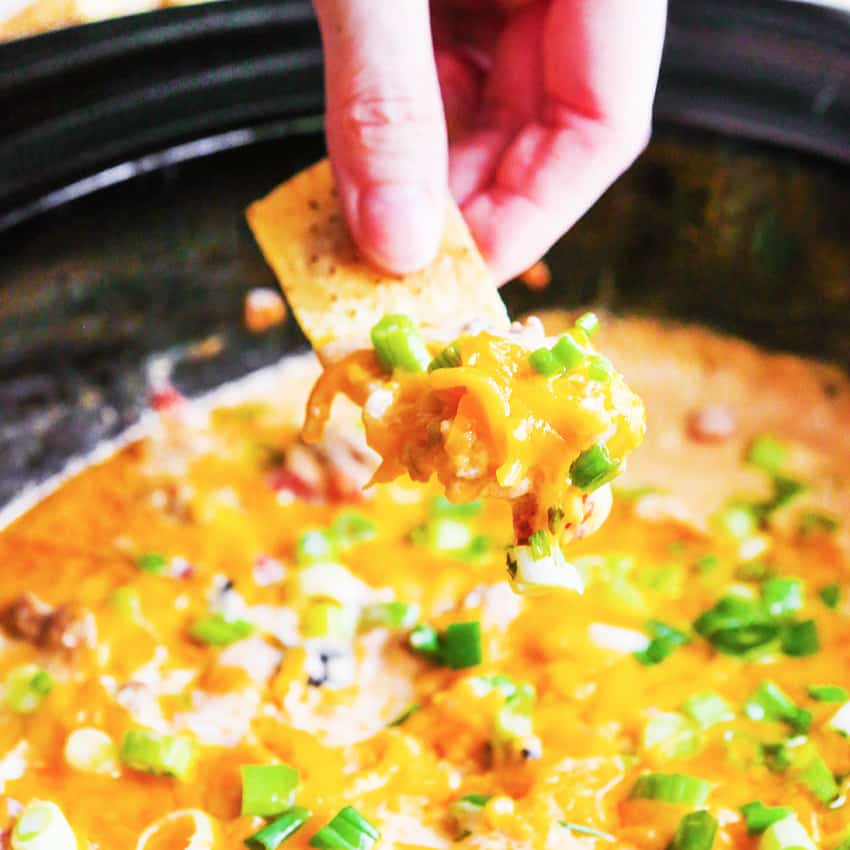 Hand holding a tortilla chip to dip into a slow cooker of sausage cheese dip. 
