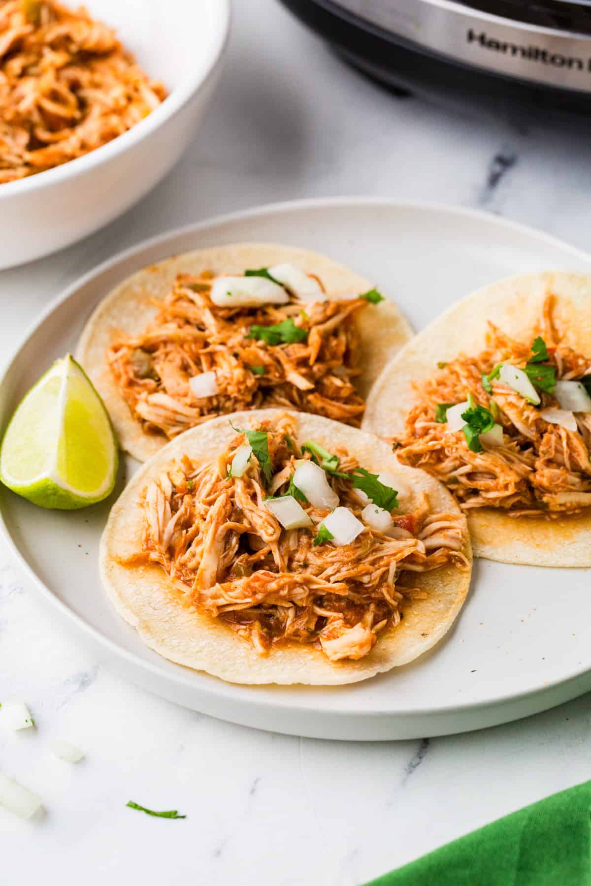 Three chicken tacos on a plate, next to a slow cooker.