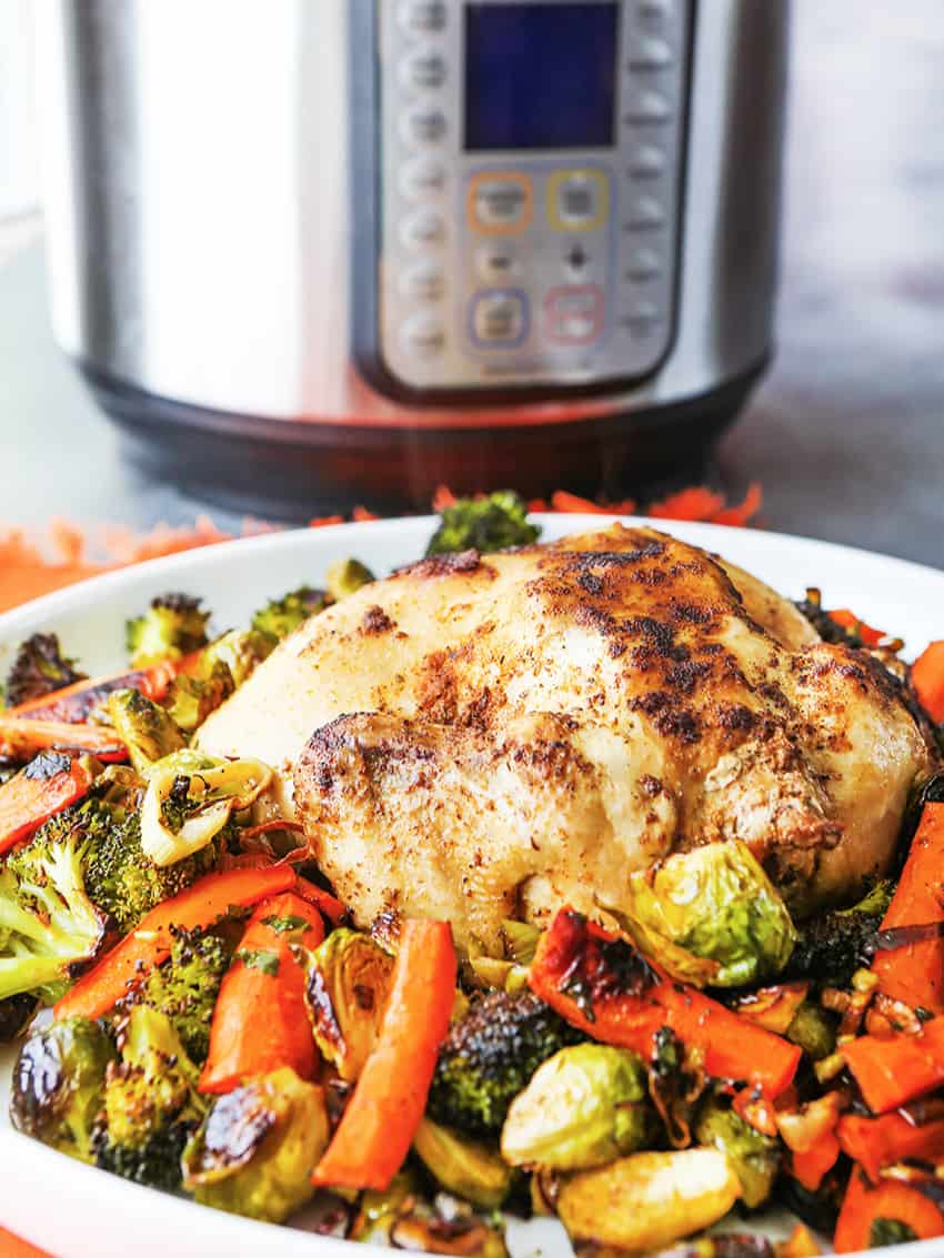 Whole chicken cooked rotisserie style on a plate surrounded with roasted vegetables. 