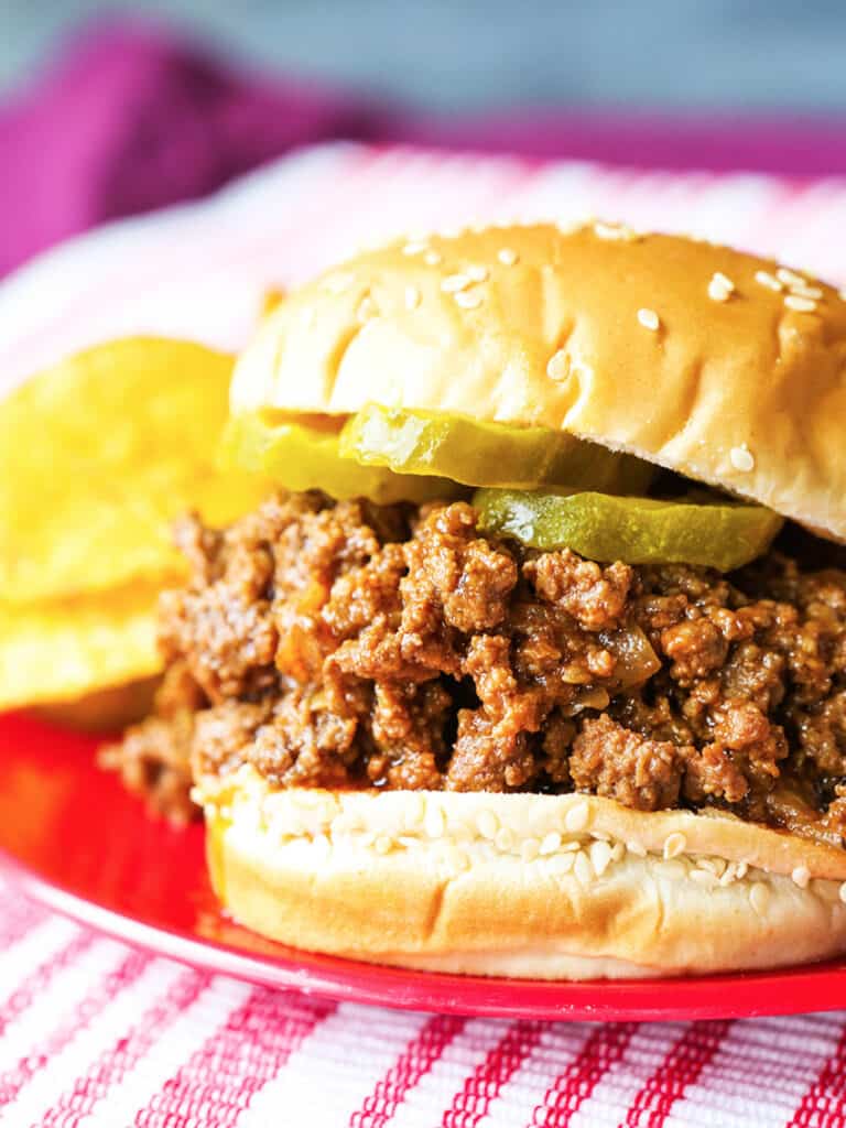 A sloppy joes sandwich on a plate with pickles placed on the meat and chips next to the sandwich. 