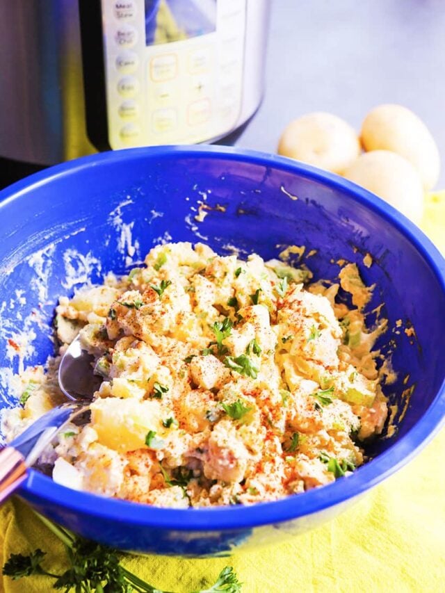 Blue bowl of instant pot potato salad with a spoon tucked into the food sitting in front of peeled hard boiled eggs and an Instant pot. 