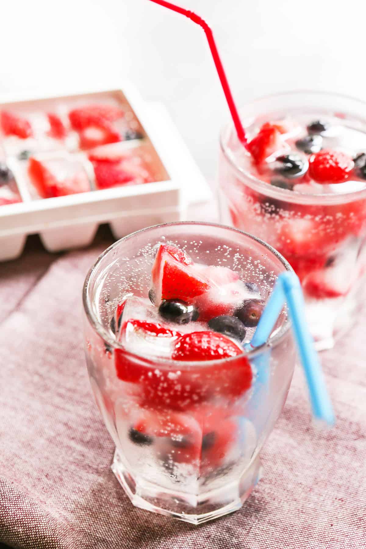 Two glasses of water with fruity cubes, next to an ice cube tray with frozen fruit cubes.