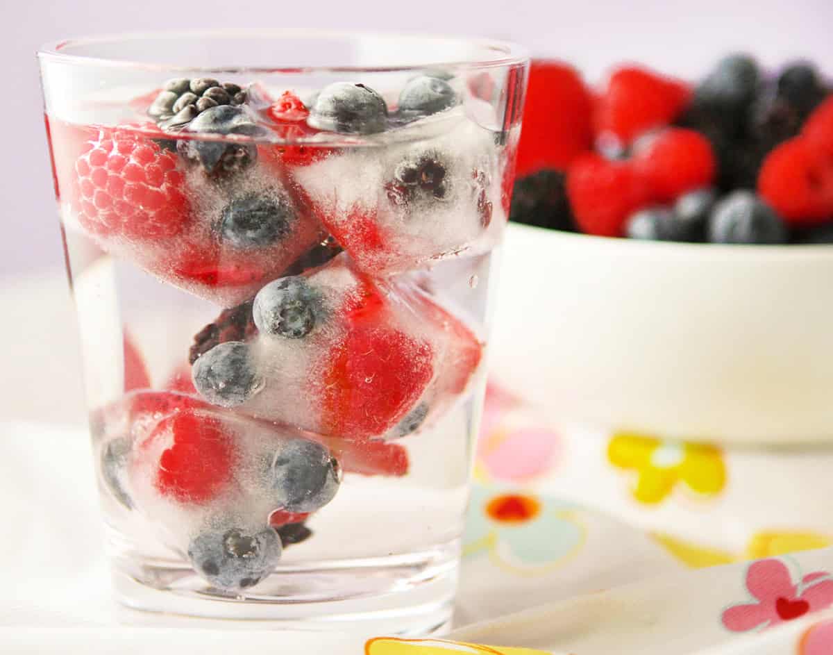 Side view of a glass of water filled with berry cubes, and a bowl of fresh berries in the background.