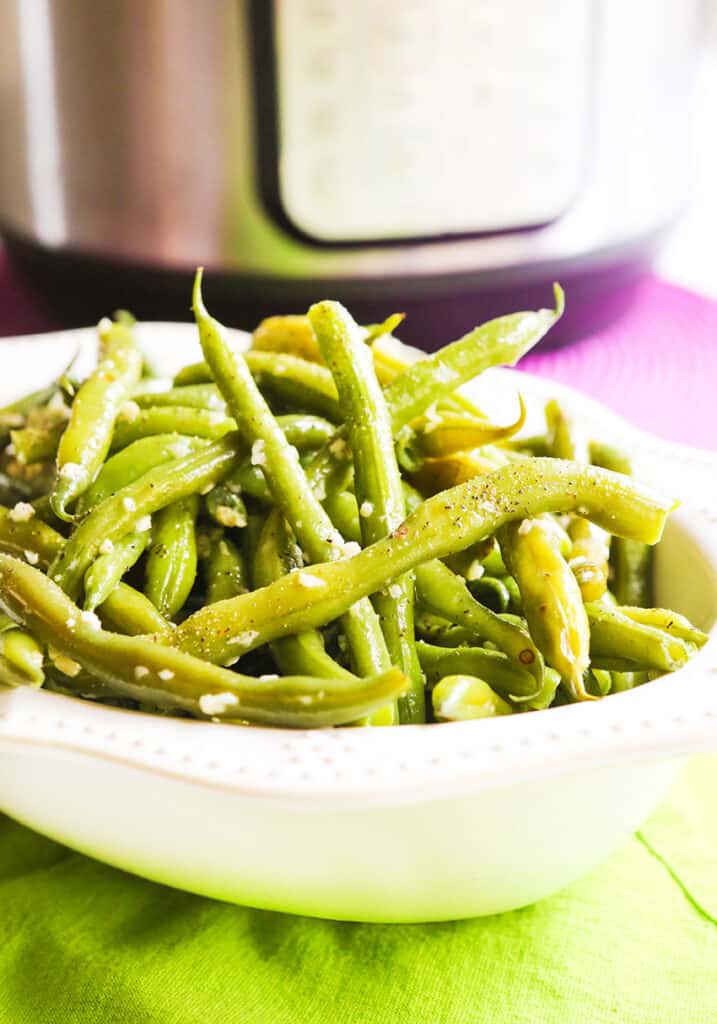 White serving bowl of green beans with sea salt sprinkled on top sitting in front of an instant pot. 