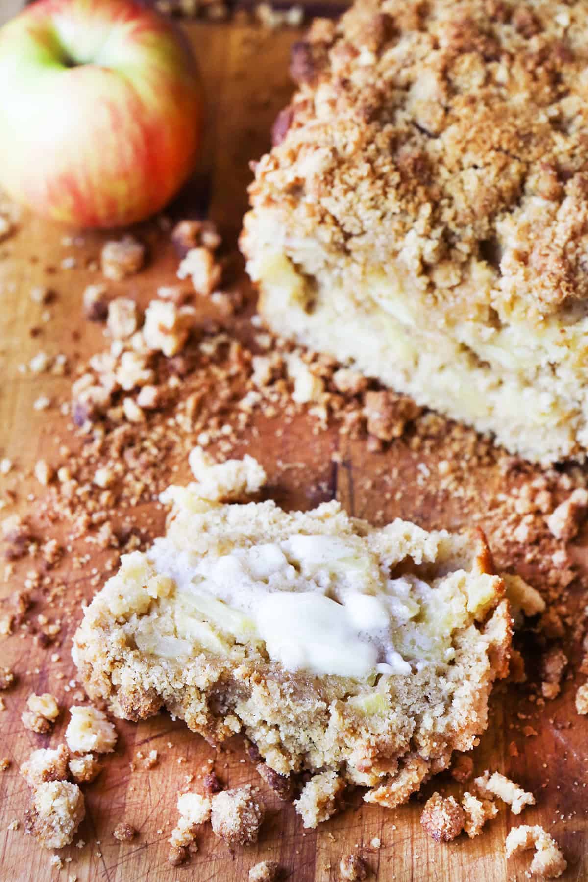 Crumbly loaf of apple bread on a cutting board, next to a slice with melted butter spread over top.