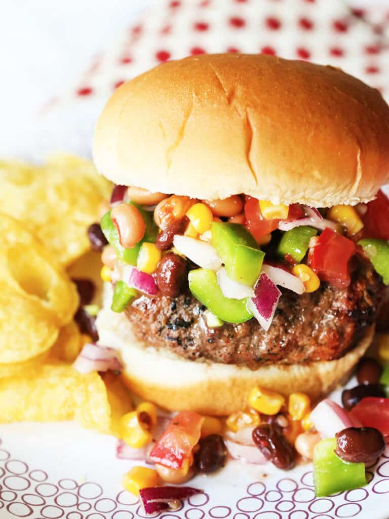 Burger with chunky salsa spilled out the edges, sitting on plate next to a pile of chips.