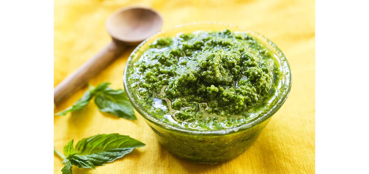 Final bowl of homemade pesto, sitting next to a few basil leaves and a spoon.