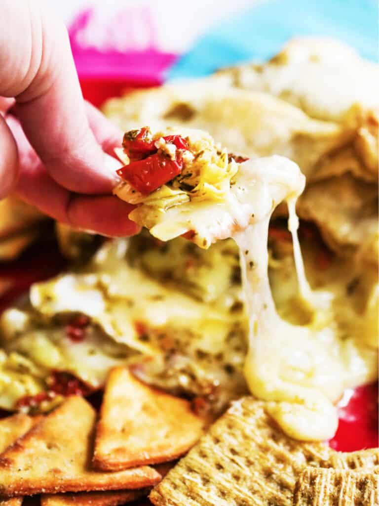 Hand scooping up baked brie packed with pesto, artichokes and sun-dried tomatoes on a cracker. 