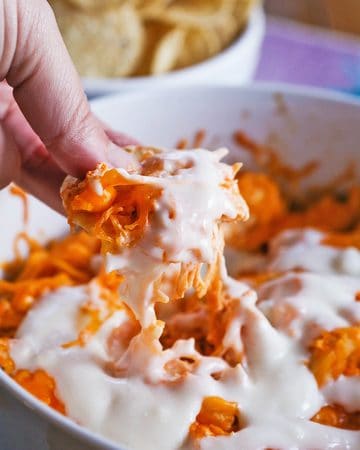 Creamy buffalo chicken dip with melted cheese in a white bowl, with a hand dipping a chip.