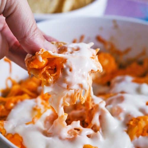 Creamy buffalo chicken dip with melted cheese in a white bowl, with a hand dipping a chip.