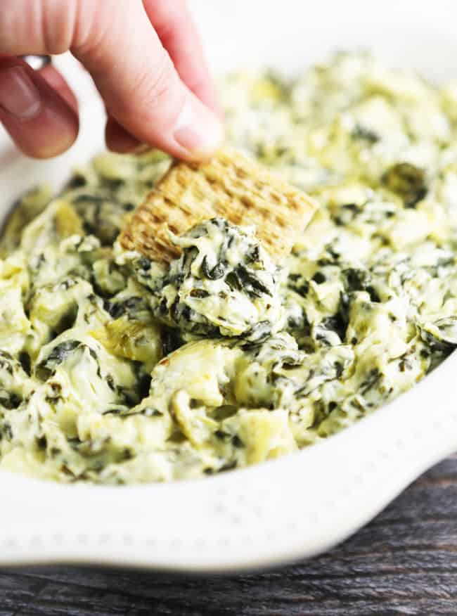 Triscuit scooping up spinach artichoke dip from a serving bowl. 