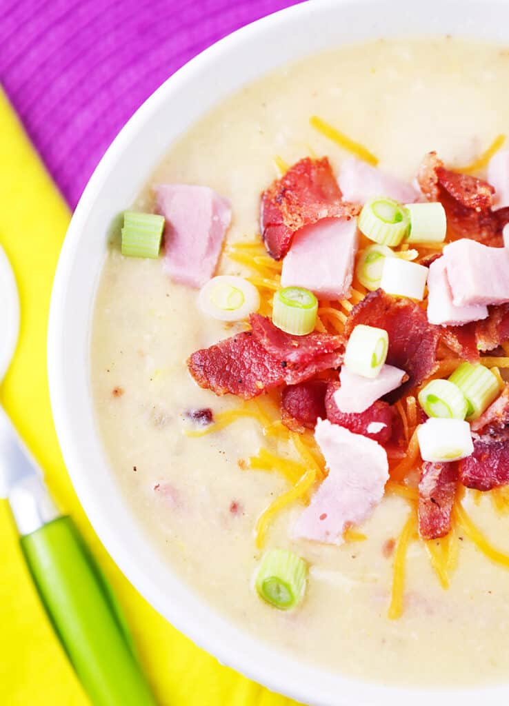 Spoon sitting next to a bowl of potato soup with chopped ham, diced bacon and green onions on top. 