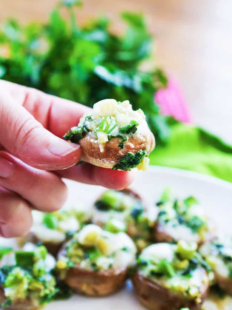 Hand holding a stuffed mushroom in front of a serving plate filled with them. 