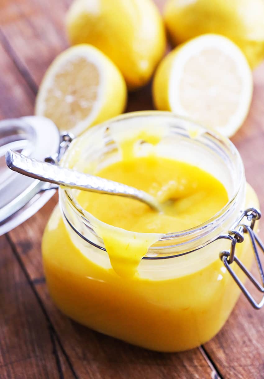 A glass jar of lemon curd with a spoon sitting inside it and fresh lemons in the background. 