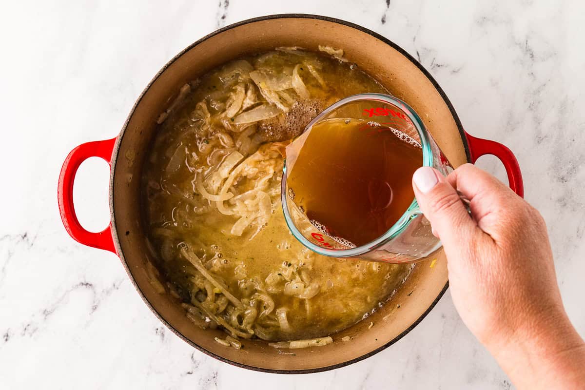 Hand pouring broth into a pan filled with caramelized onions.