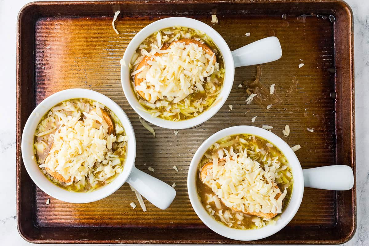 Three bowls of french onion soup on a baking sheet ready to melt cheeses in oven.