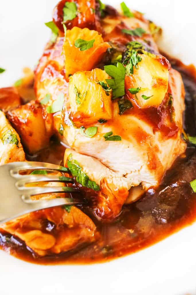 Fork cutting into chicken breast covered in sauce and chunks of pineapple on a plate. 