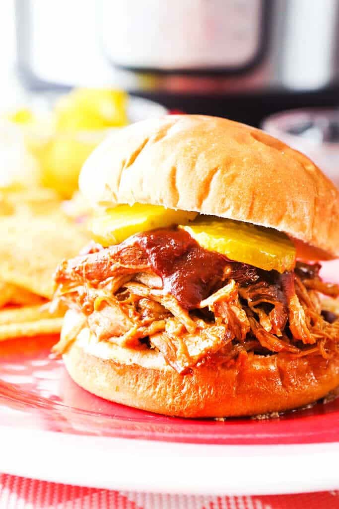 Pulled pork sandwich topped with pickled and bbq sauce sitting on a plate in front of the instant pot. 