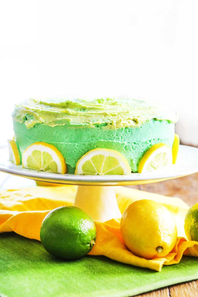 lemon layer cake on cake stand with lemons and limes beneath it