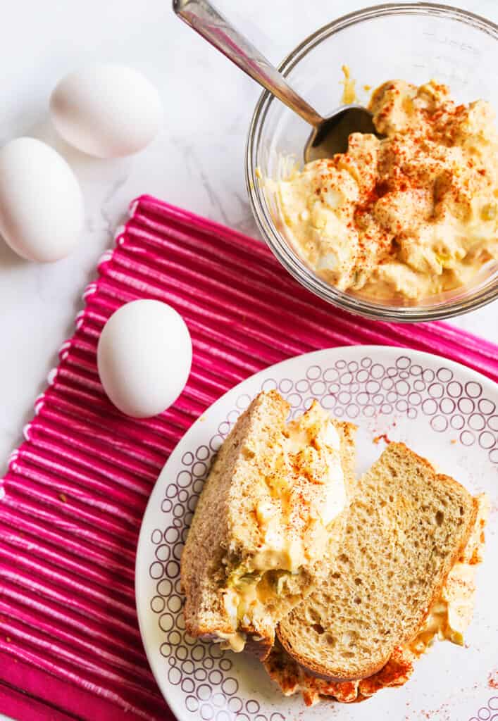 top view of an egg salad sandwich on plate next to bowl of the salad mixture