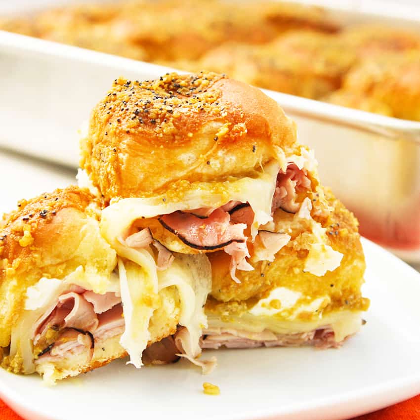 Ham and cheese sliders stacked on one another on a plate with cheese pulls coming out of them.