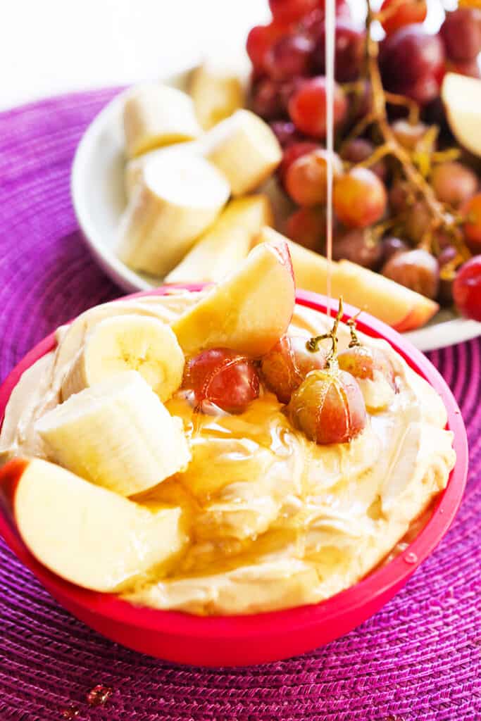 Honey being drizzled over healthy frit dip that's garnished with grapes, apple slices and banana chunks. 