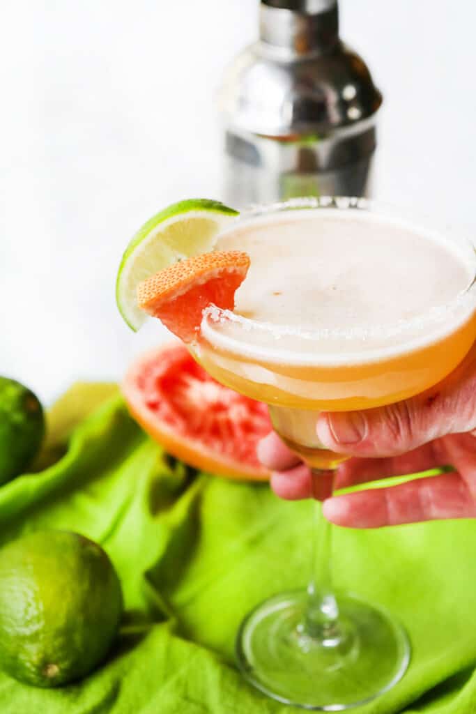 hand holding out a grapefruit margarita with a garnish of lime and grapefruit slice