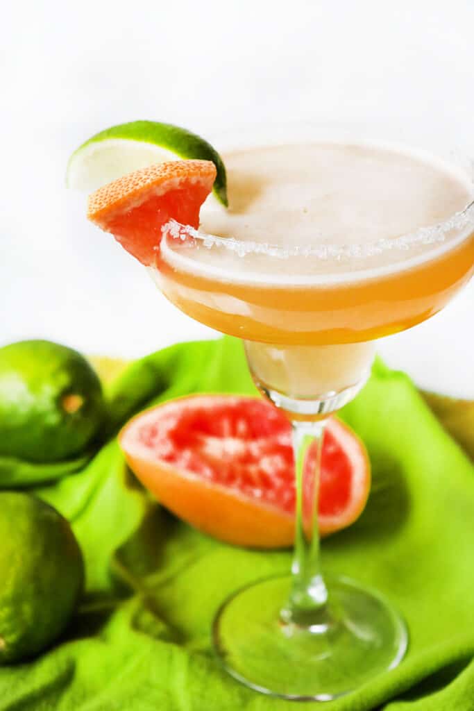 A graperfruit margarita with limes and squeezed grapefruit lying besides it. 