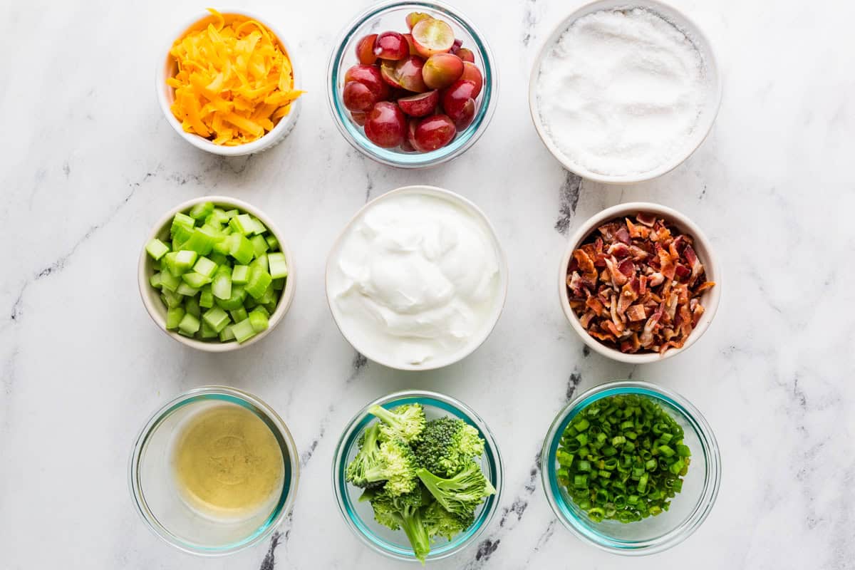 Broccoli salad ingredients in individual bowls on a white counter.