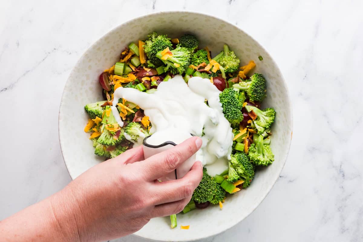 Hand pouring dressing over broccoli salad in a mixing bowl.