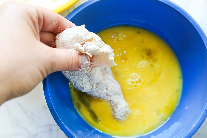hand dipping flour covered piece of chicken into beaten eggs
